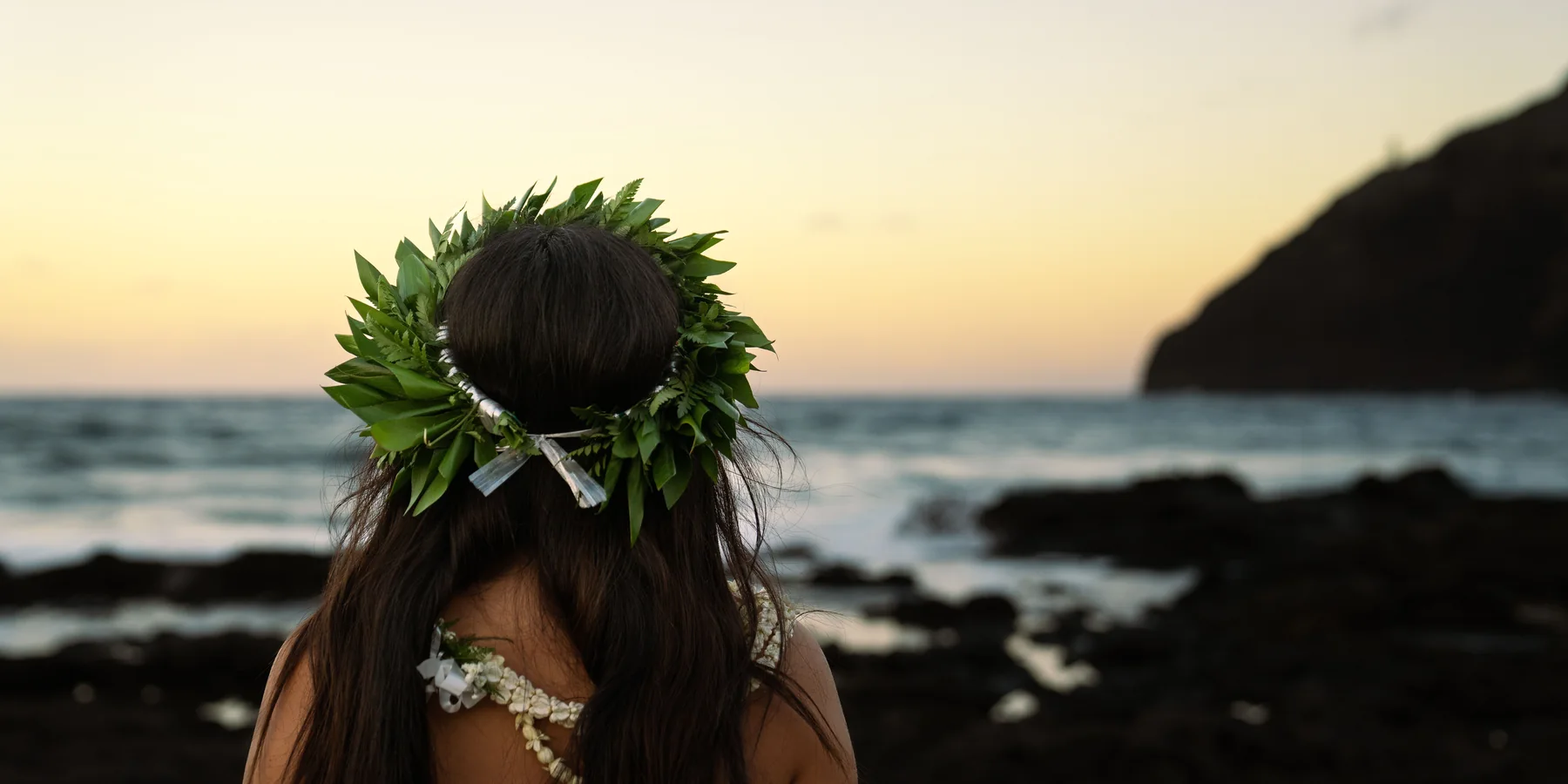 A woman's green ti-leaf lei seen from behind, silhouetted against the ocean and a dark headland at dusk